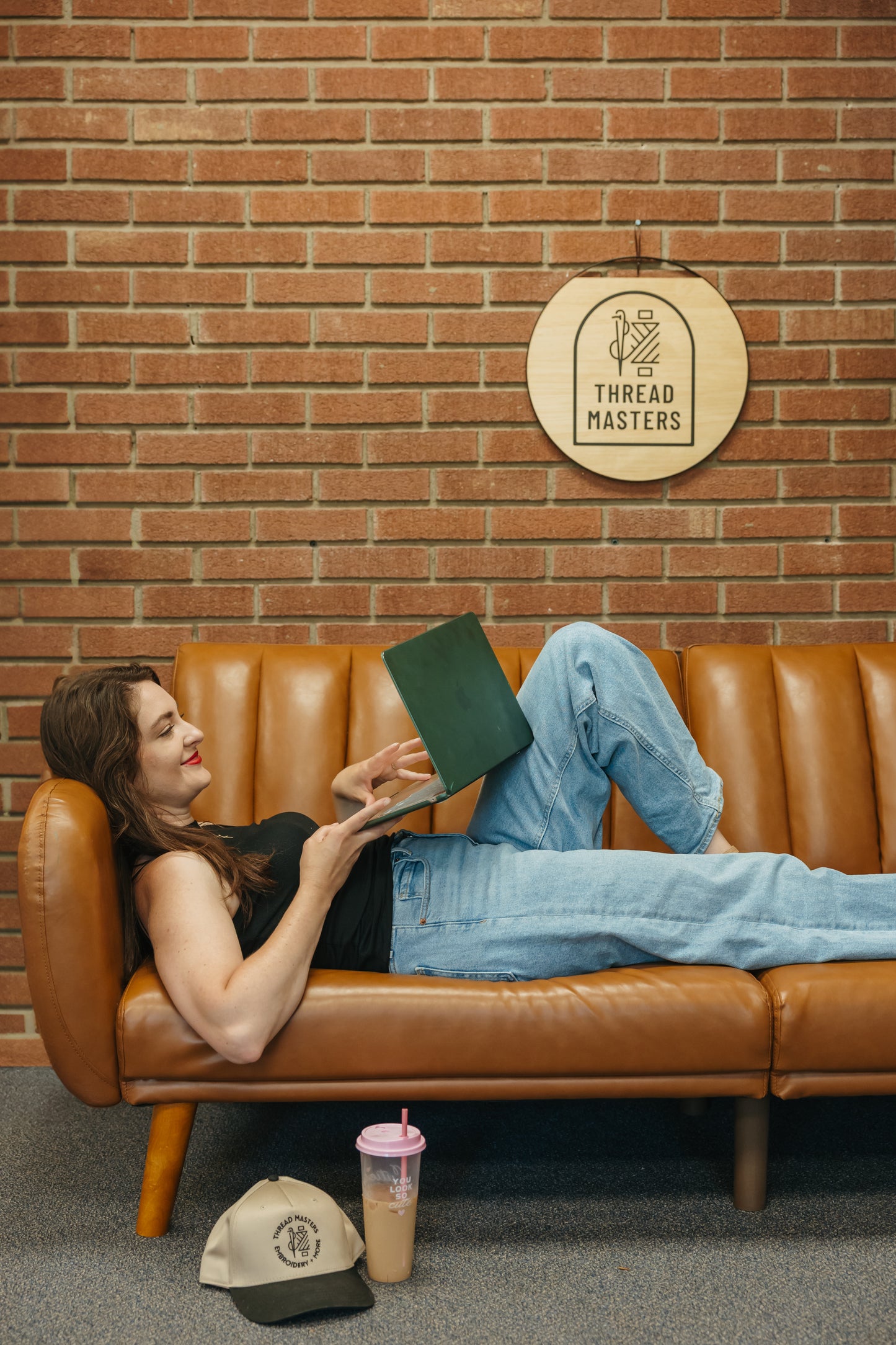 Woman lying on a brown leather couch with a green book, wearing a black top and blue jeans, in front of a brick wall with 'Thread Masters' sign.