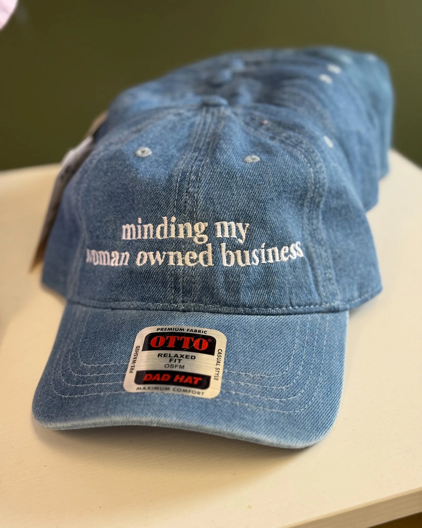 Blue denim cap with 'minding my woman owned business' text on a beige surface.