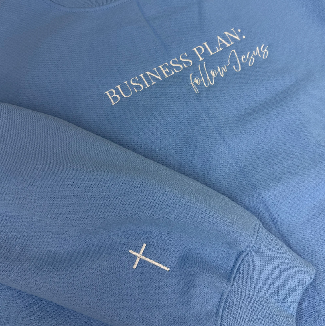 Blue sweatshirt with 'Business Plan: Follow Jesus' text and cross logo.
