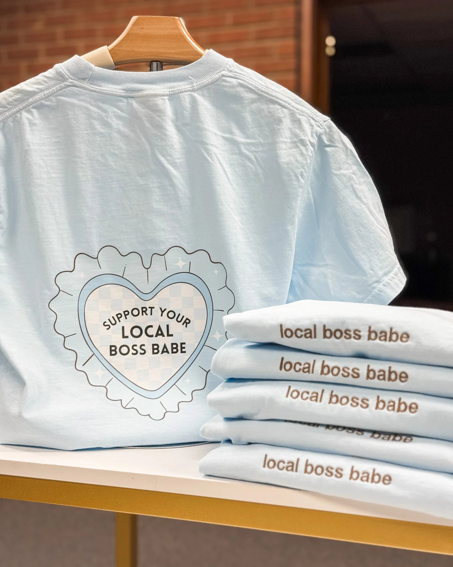Light blue t-shirt with 'Support Your Local Boss Babe' text on a wooden hanger, with more shirts stacked below.